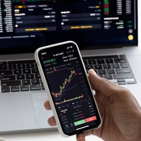 Person analyzing stock market trends on smartphone with laptop background
